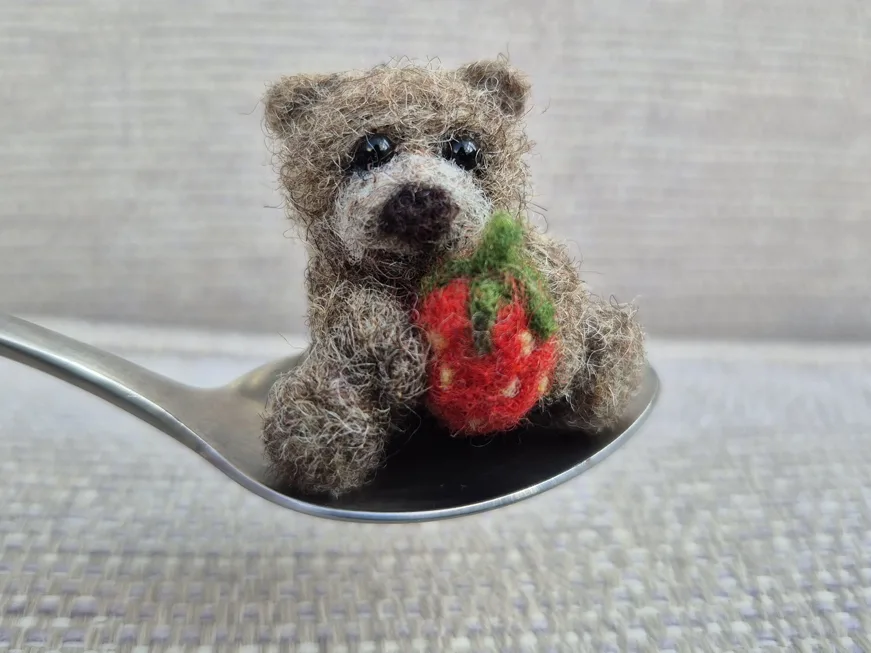 A tiny felted bear holding a strawberry, sitting in a teaspoon