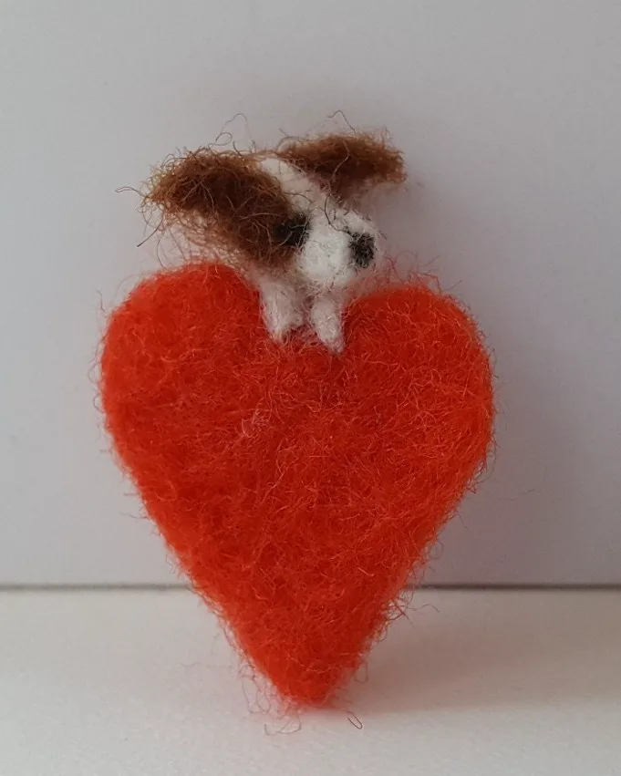 A tiny needle-felted dog peeping over a red felted heart, made by Grace