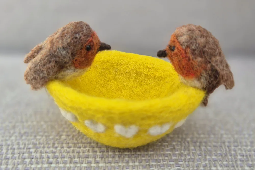 Two needle-felted robins perched on the rim of a yellow felted ring bowl